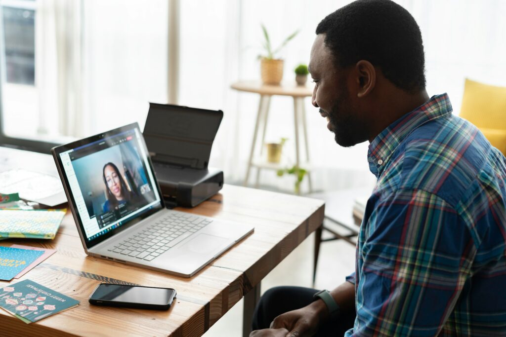 Man taking a sales call on a laptop on his kitchen table the man is waring a blue button up shirt and watch.