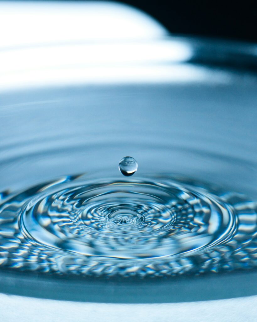 Close-up of a single water droplet suspended above rippling water, symbolizing purity, clarity, and the ripple effect of positive change—reflective of the Darren and Mike Enagic wellness mission