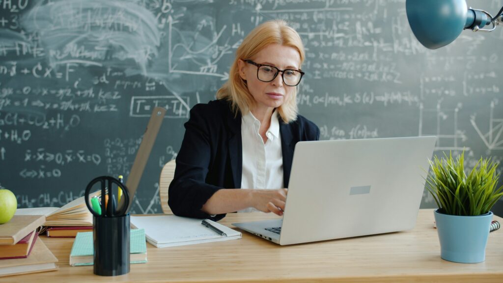 Teacher in glasses working on a laptop at her desk by a chalkboard, building online income as a side hustle for educators