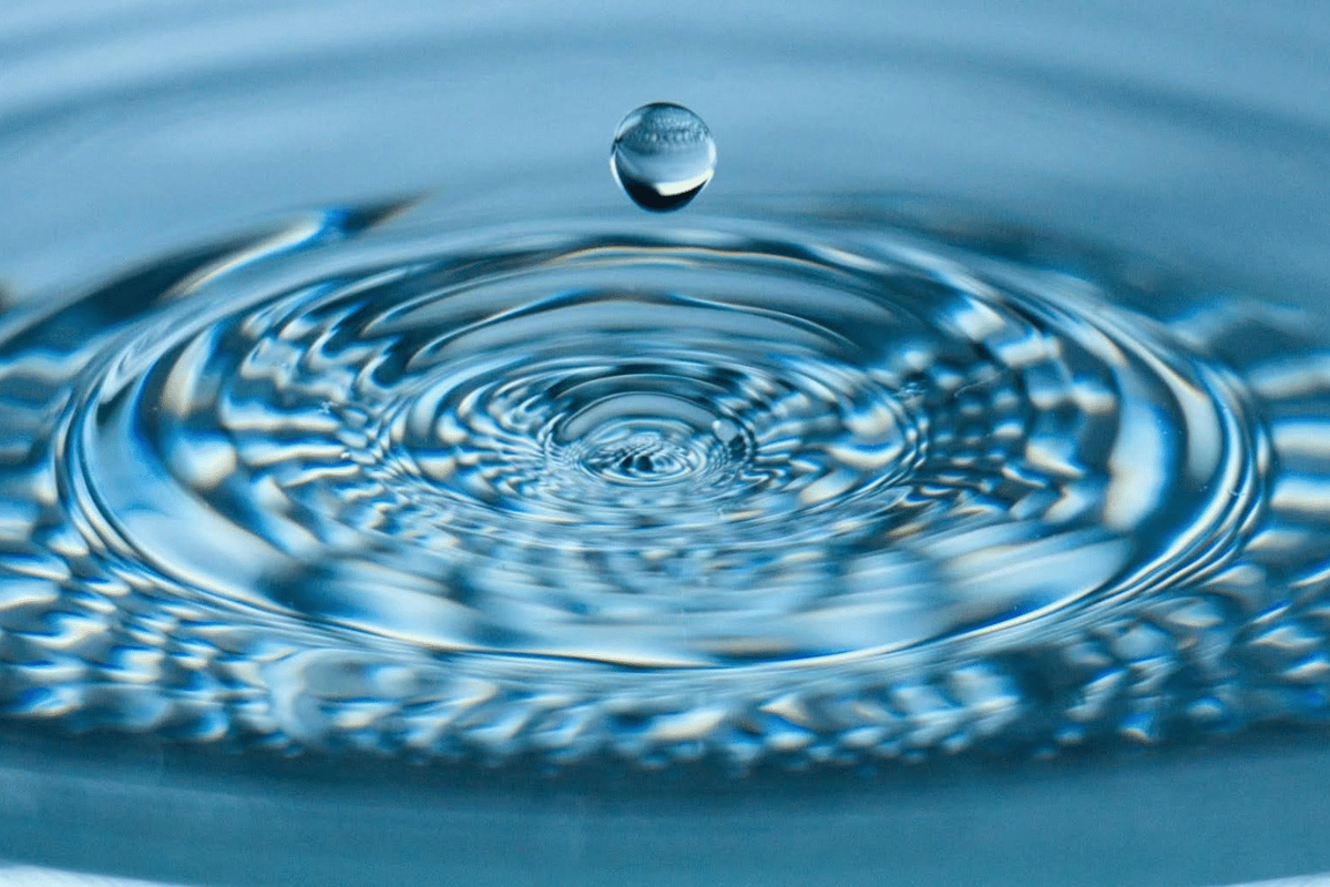 Close-up of a single water droplet suspended above rippling water, symbolizing purity, clarity, and the ripple effect of positive change—reflective of the Darren and Mike Enagic wellness mission.