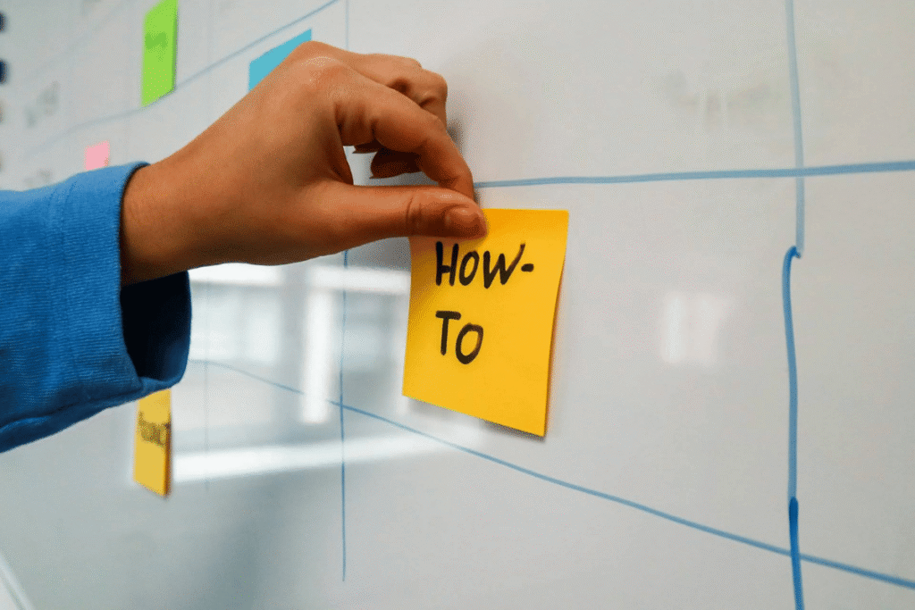 Person placing a yellow sticky note labeled "HOW-TO" on a whiteboard, symbolizing step-by-step planning or instructional guidance for starting a digital business.